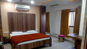 A modern hotel room with a wooden bed, an air conditioning unit, a small table, and a window with curtains. - Hotel Tak (Udaipur)