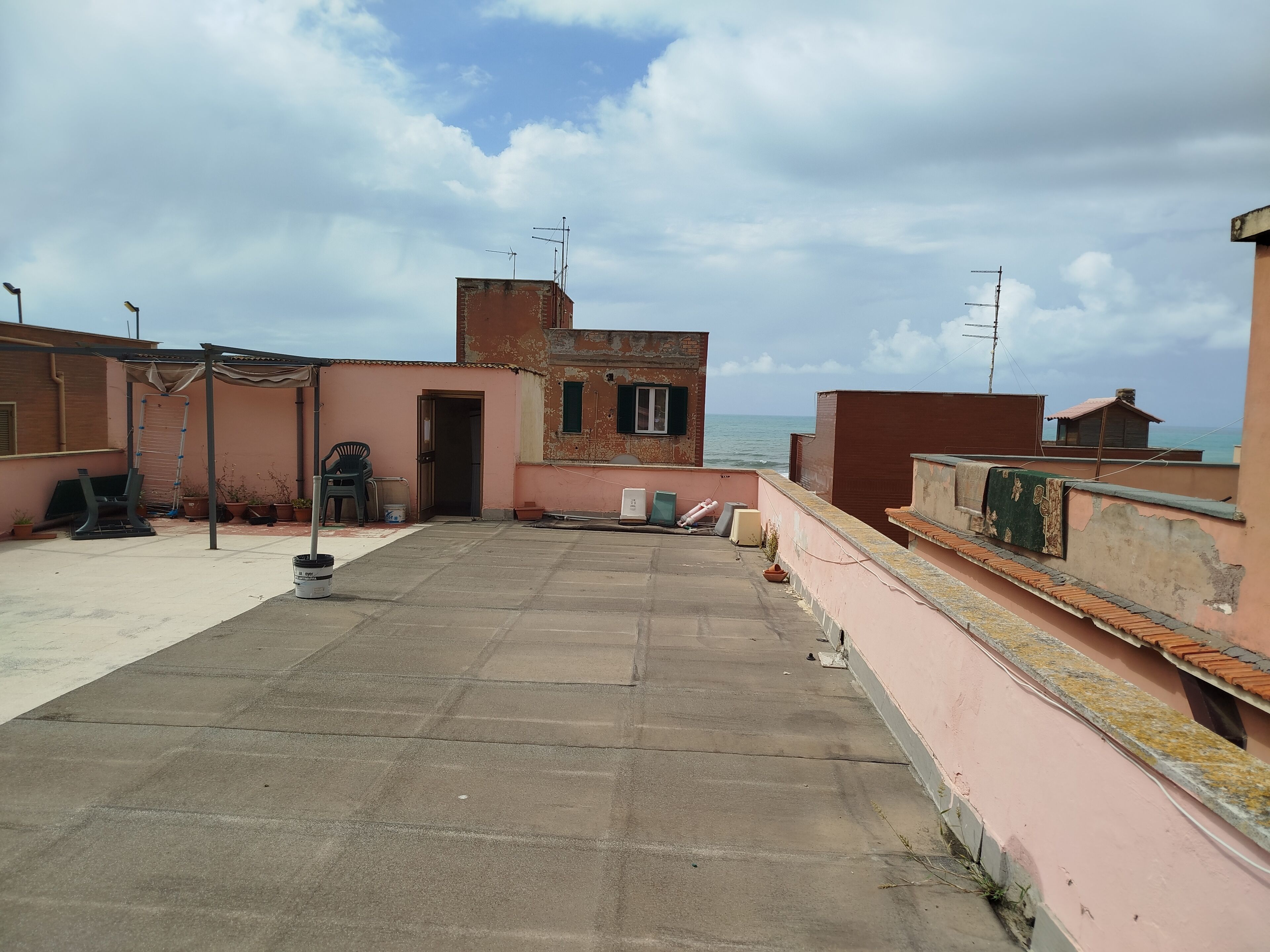 A rooftop terrace with a view of the sea, featuring a small building with a door and windows, and a concrete floor.