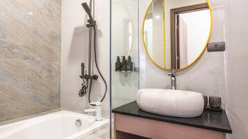 A modern bathroom with a glass-enclosed shower, a white bathtub, a round mirror with a gold frame, and a wooden vanity with a black countertop.