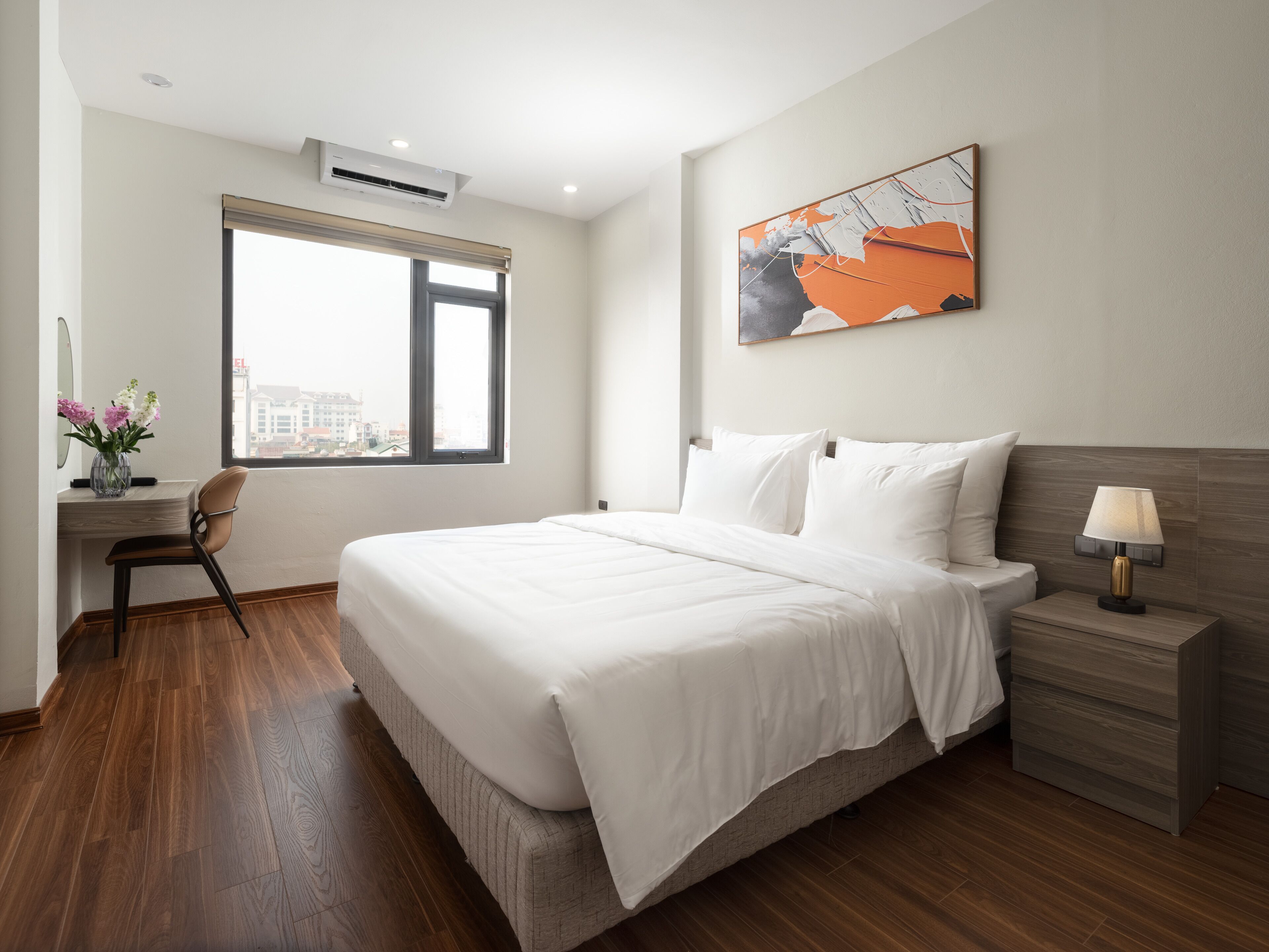 A modern hotel room with a large bed, a desk with a chair, a nightstand, and a window with a view of buildings.