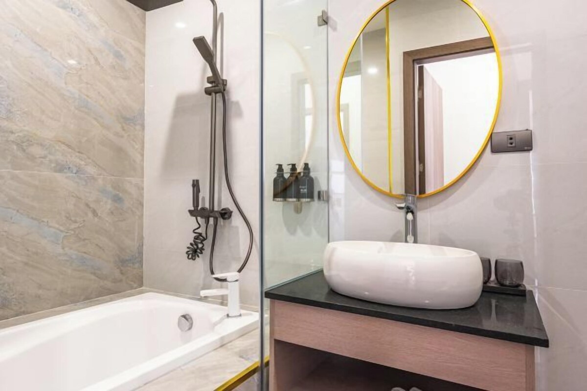 Un baño moderno con ducha de vidrio, un lavamanos blanco y un espejo redondo con marco dorado.