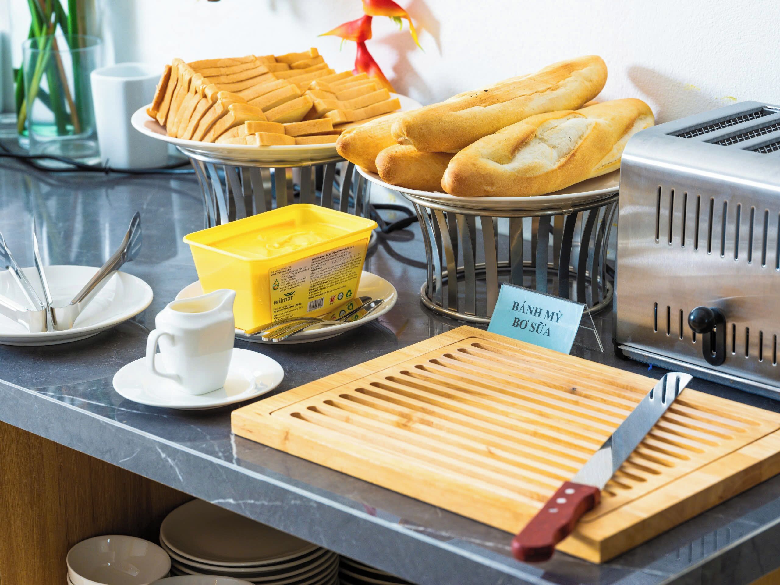 A buffet setup with various food items, a sign indicating 'BÁNH MY BỘ SỬA', and a toaster.