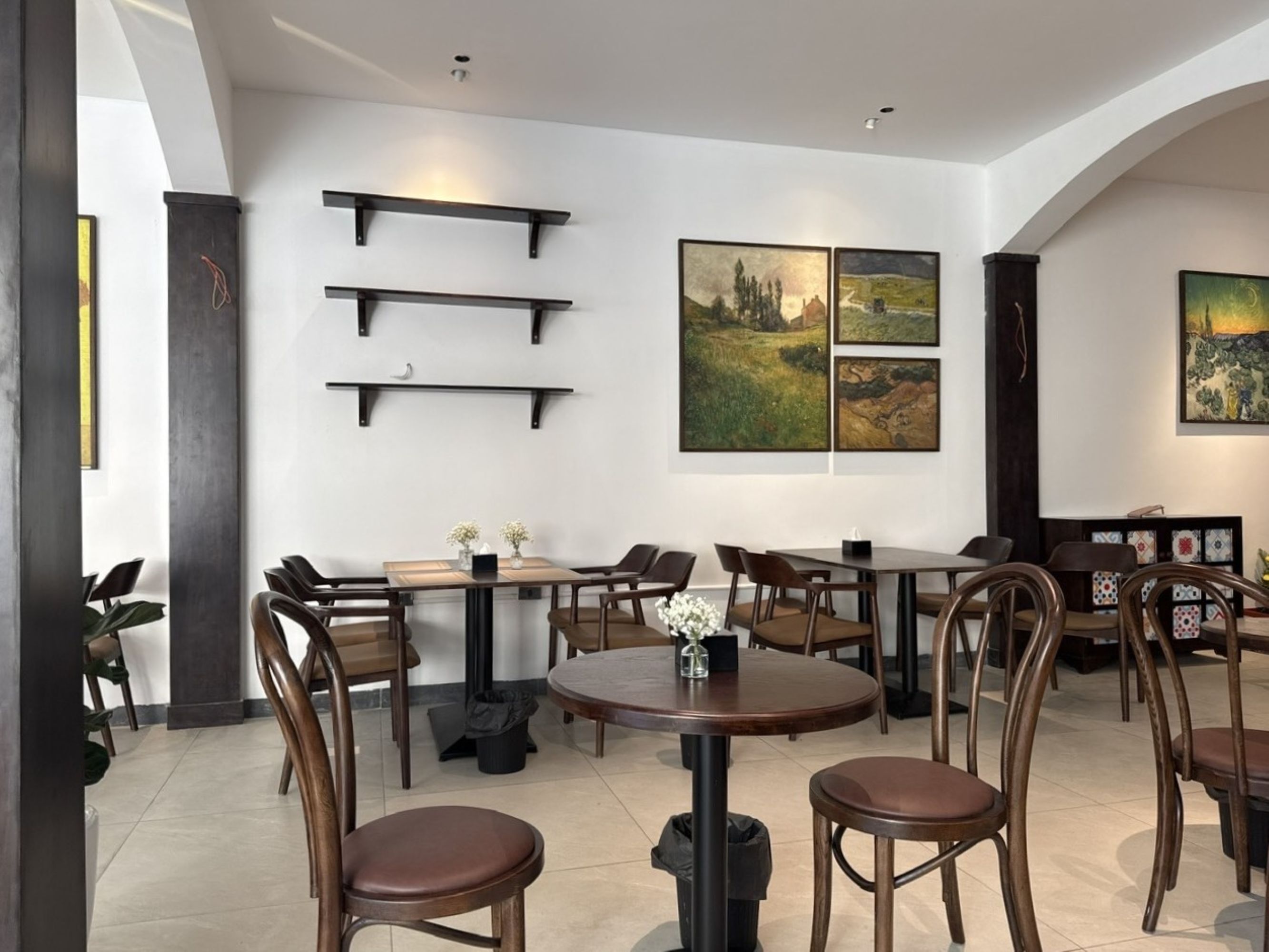 A modern dining area with multiple tables and chairs, black wall-mounted shelves, and framed landscape paintings.