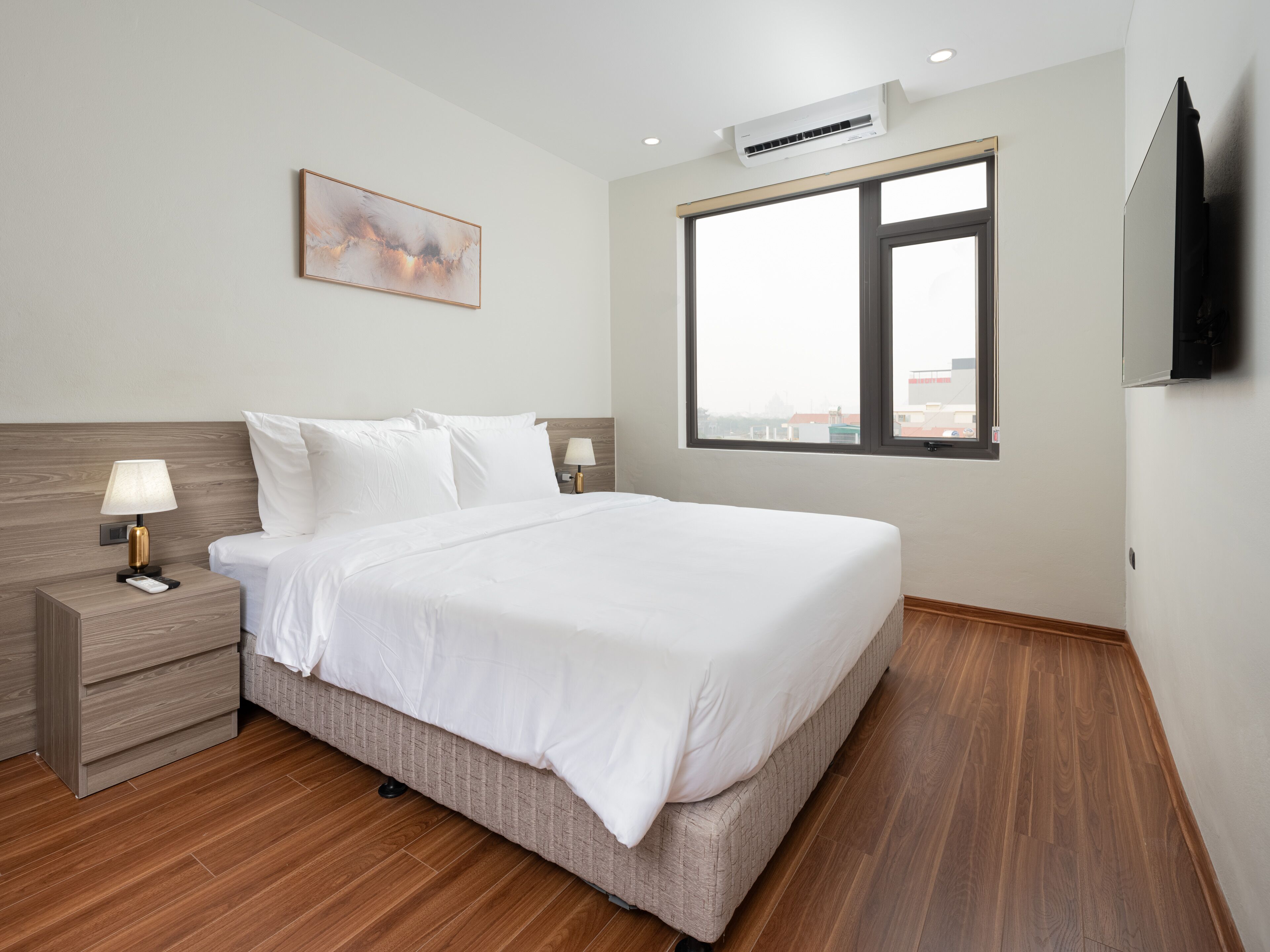 A modern hotel room with a large bed, a bedside table with a lamp, an air conditioning unit, a large window, and a painting on the wall.