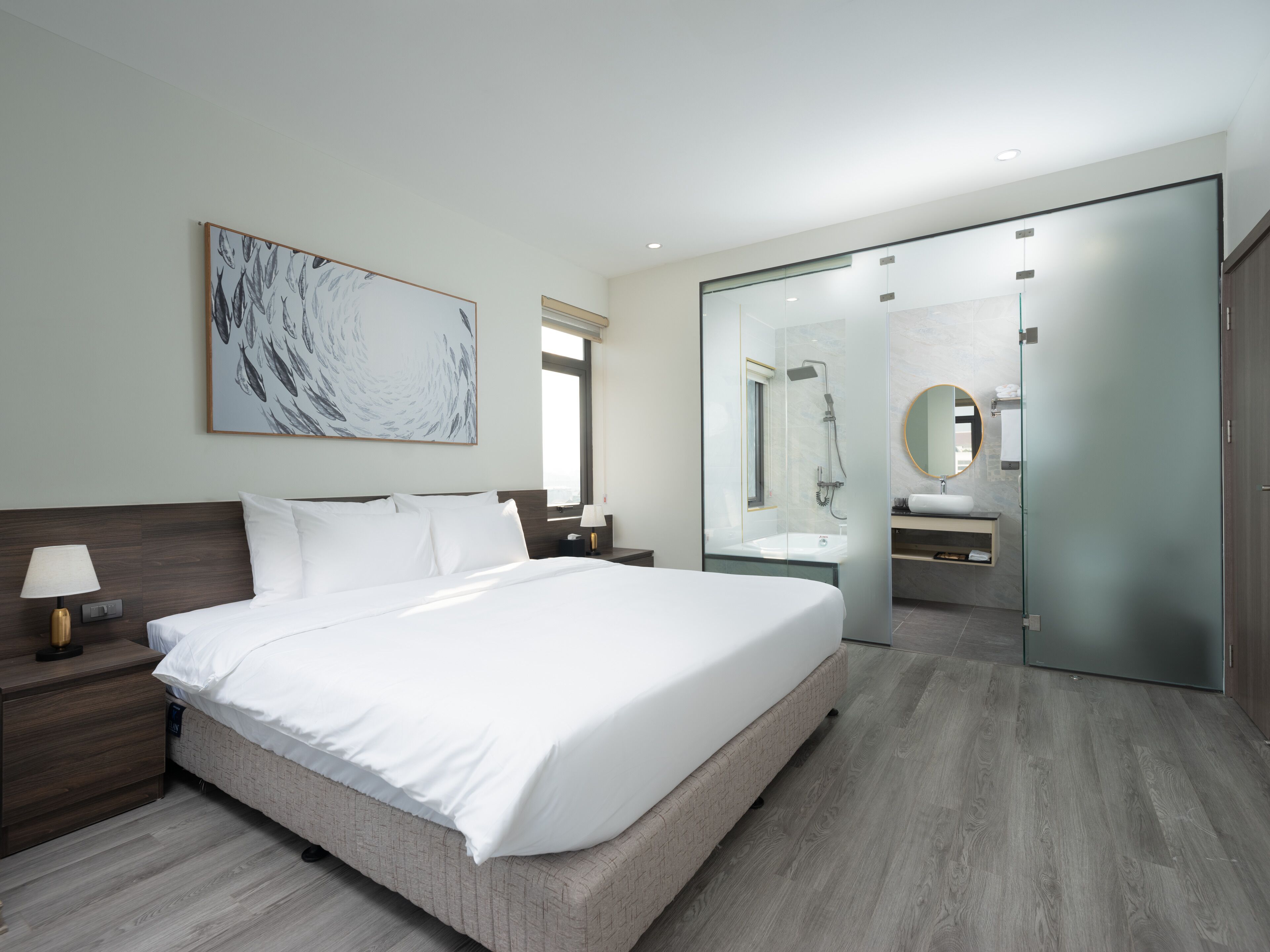 A modern hotel room with a large bed, a nightstand, a bedside table with a lamp, a large abstract painting on the wall, a glass-enclosed bathroom with a mirror, and a wooden floor.