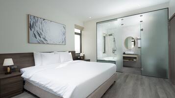 A modern hotel room with a large bed, a nightstand, a bedside table with a lamp, a large abstract painting on the wall, a glass-enclosed bathroom with a mirror, and a wooden floor.