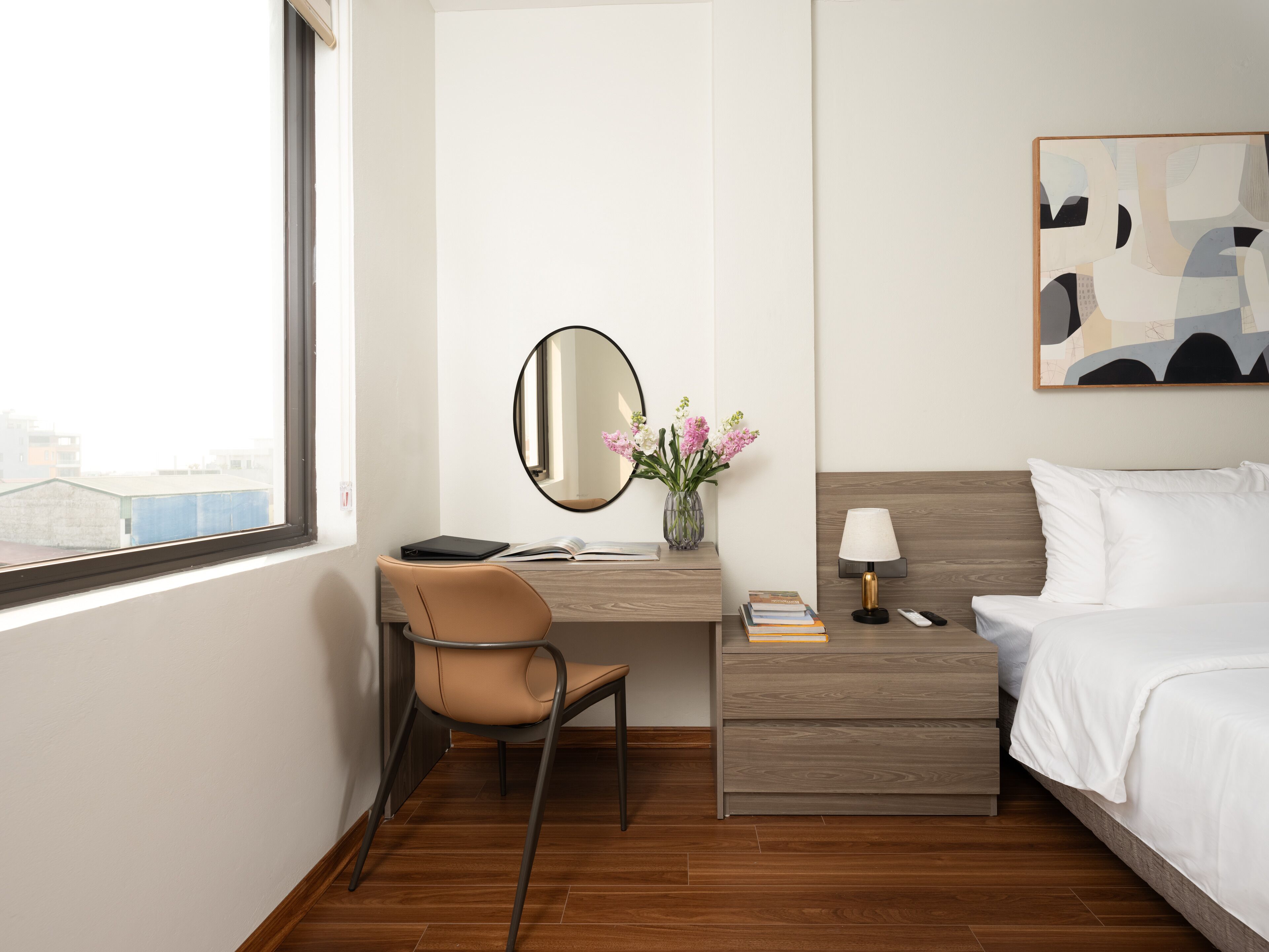 A hotel room with a desk, chair, bedside table, lamp, and a bed.