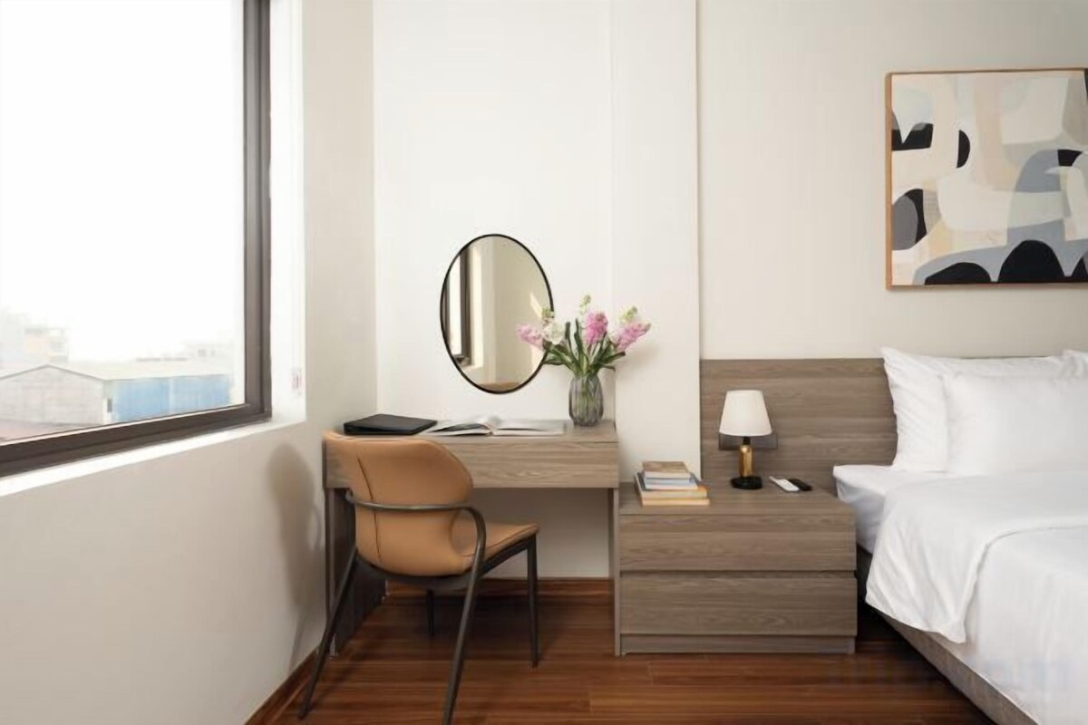 A hotel room with a bed, a desk with a chair, a mirror, a vase with flowers, and a painting on the wall.