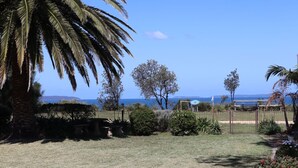 A garden with a view of the sea, featuring palm trees, a stone table, and a wooden fence.
