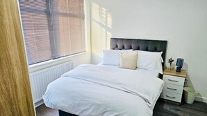 A bedroom with a bed, bedside table, and a window with blinds. - Delightful Double En-suite Room (Ponders End)