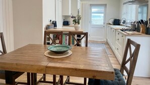 A modern kitchen with a wooden dining table, white cabinets, and a view of the sea through a large window.