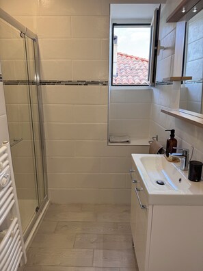 A modern bathroom with a glass shower enclosure, a white sink with a mirror above it, and a window providing natural light. - Apartment in the heart of the village (murato)