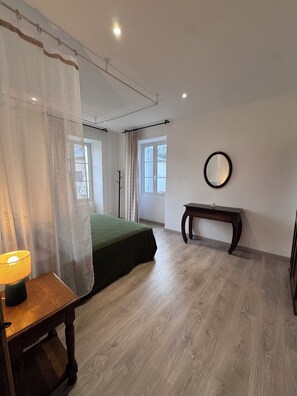 A bedroom with a bed, a bedside table with a lamp, a mirror on the wall, and a window with curtains. - Apartment in the heart of the village (murato)