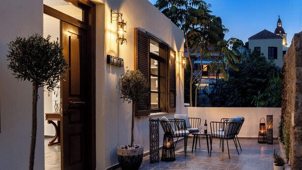 A well-lit outdoor patio with a table and chairs, surrounded by potted plants and a stone floor. - Alegrand Suites (Rhodes)