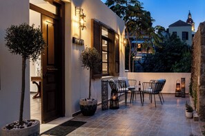 A well-lit outdoor patio with a table and chairs, surrounded by potted plants and a stone floor. - Alegrand Suites (Rhodes)