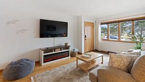 A modern living room with a flat-screen TV, wooden entertainment unit, and a large window offering a view of the outdoors.