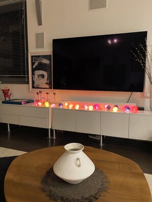 Un salon moderne avec un téléviseur à écran plat, un meuble blanc, des guirlandes lumineuses colorées et un vase décoratif posé sur une table en bois.