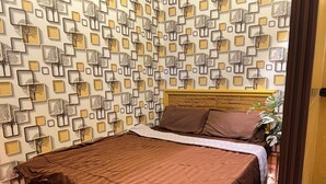 Ein ordentlich bezogenes Bett mit brauner Tagesdecke, ein Kopfteil mit Holzoptik und eine Wand mit geometrischen Formen und Bäumchen-Motiven.