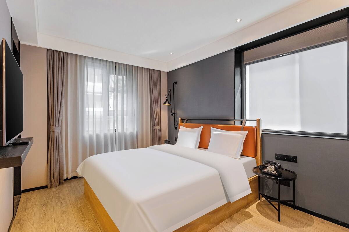 A modern hotel room with a large bed, a television on a stand, a small table with a telephone, and a window with curtains.