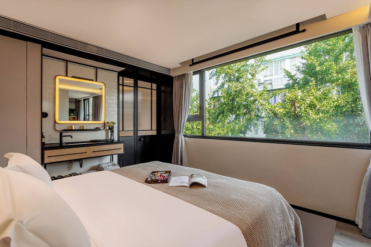 A modern hotel room with a large bed, a bedside table with a book and a magazine, a mirror with a light, and a window with a view of trees and buildings.