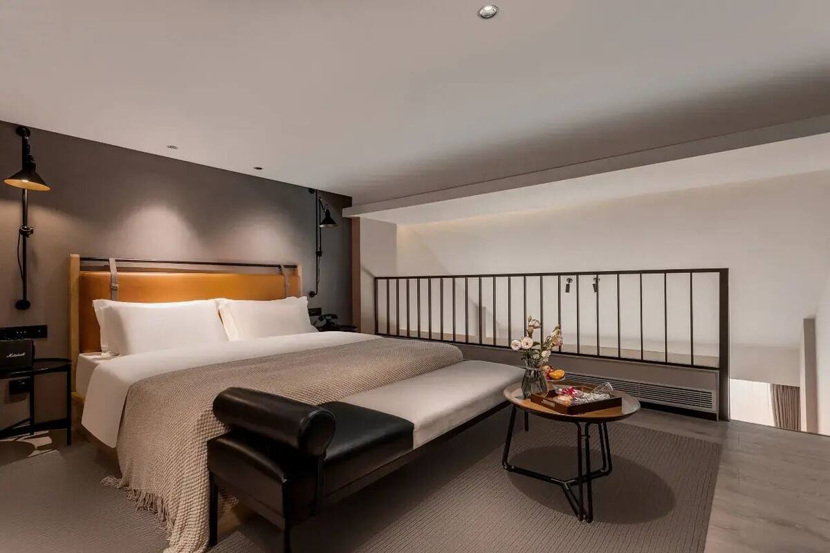A modern hotel room with a large bed, a bedside table with a vase of flowers, a small round table with a tray, and a view of the city through a large window.