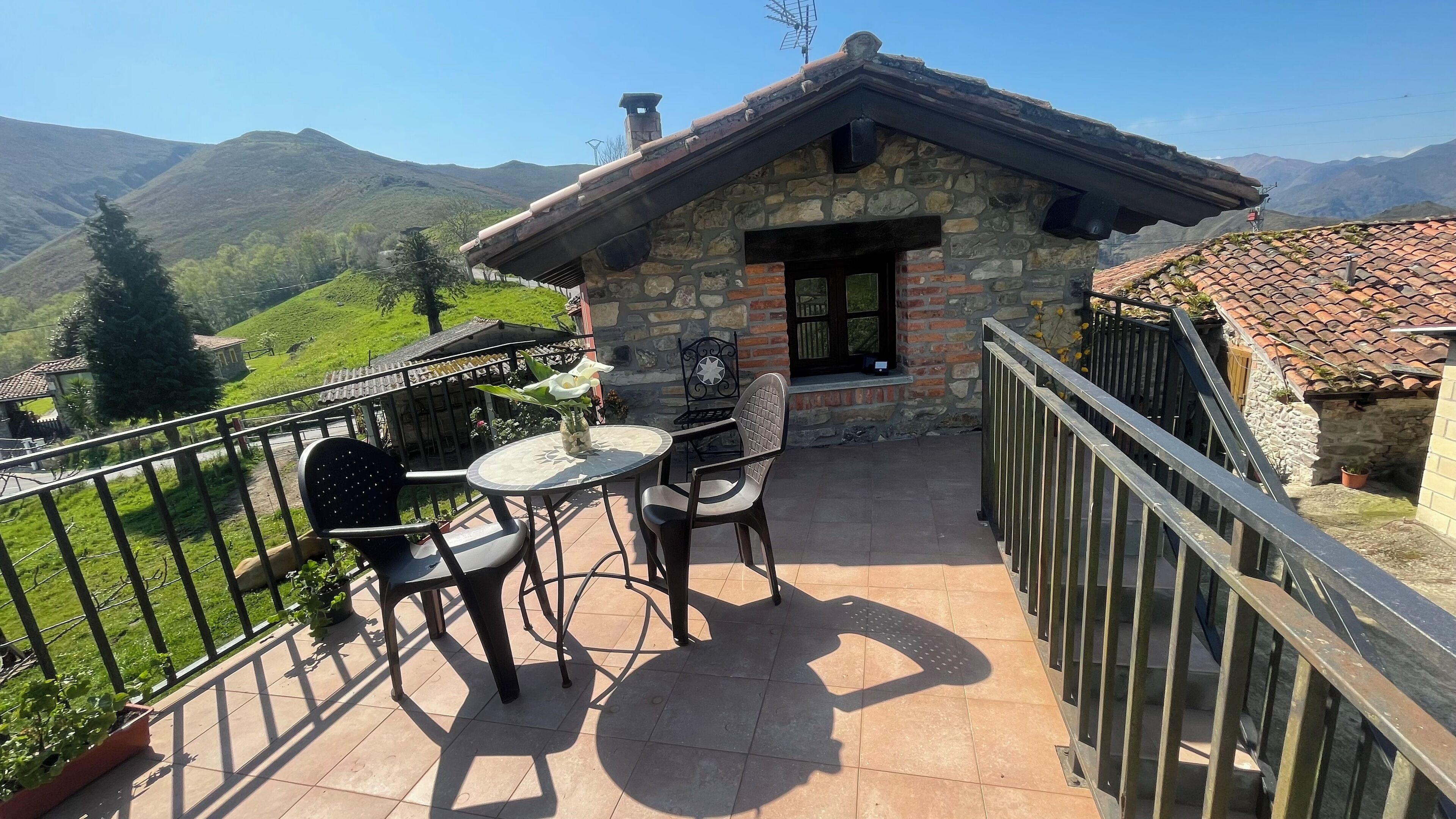 Un edificio in pietra con tetto in tegole, un balcone con tavolo e sedie e vista su dolci colline e montagne.