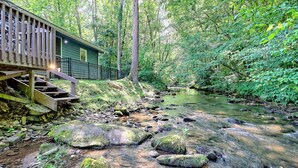 Una cabaña con terraza de madera y escaleras que conducen a un porche, rodeada de un bosque y un arroyo.