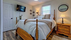 Un dormitorio con una cama de madera, un ventilador de techo, un televisor, una lámpara de noche, una ventana y un espejo.