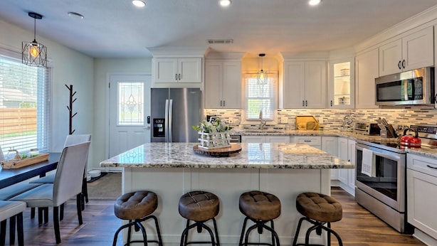 Una cocina moderna con armarios blancos, una isla grande con taburetes de bar, electrodomésticos de acero inoxidable y un centro de mesa con planta decorativa.