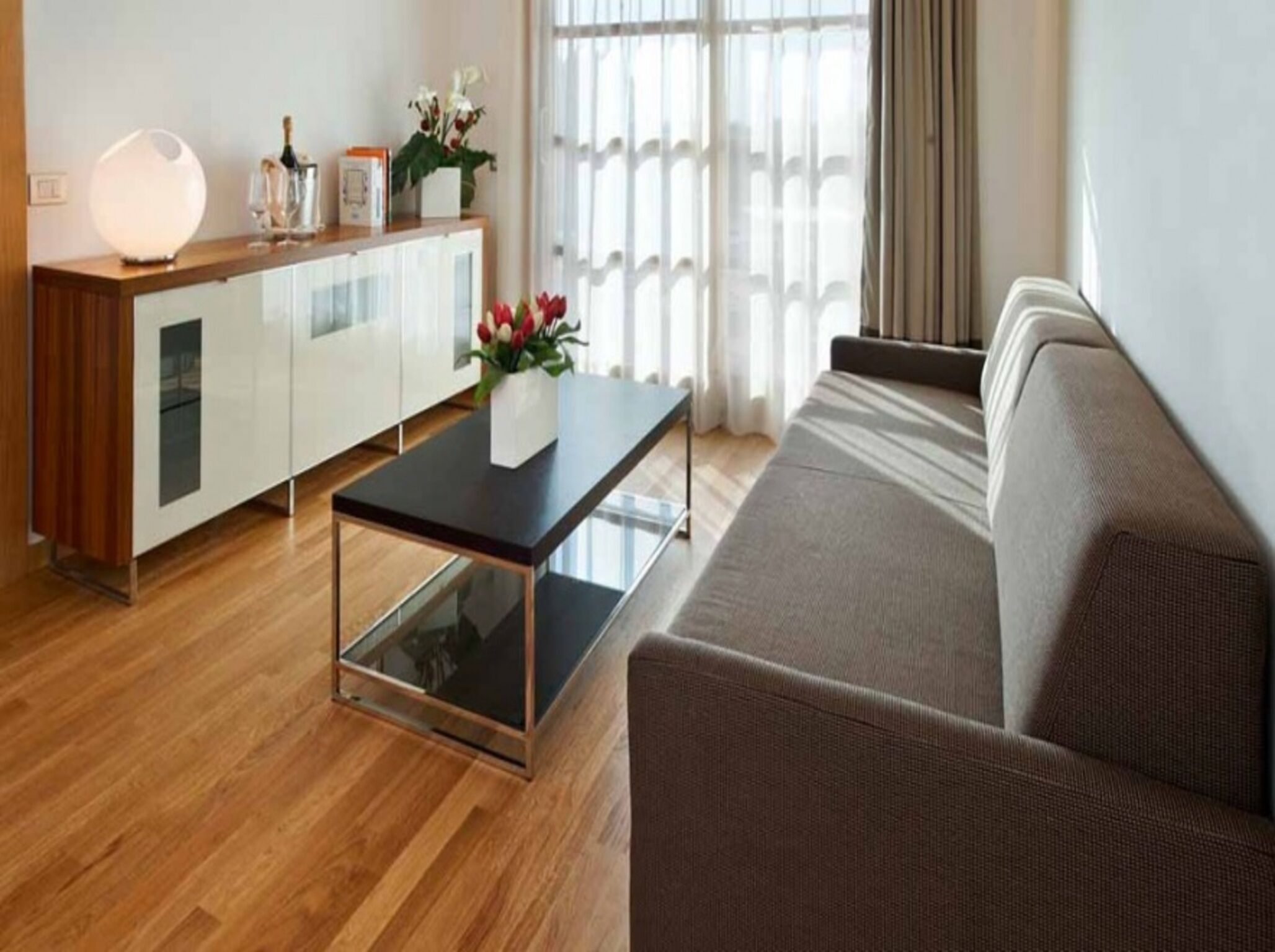 Una sala de estar moderna con piso de madera, una mesa de centro de vidrio, un sofá gris y un armario blanco con puertas de cristal.