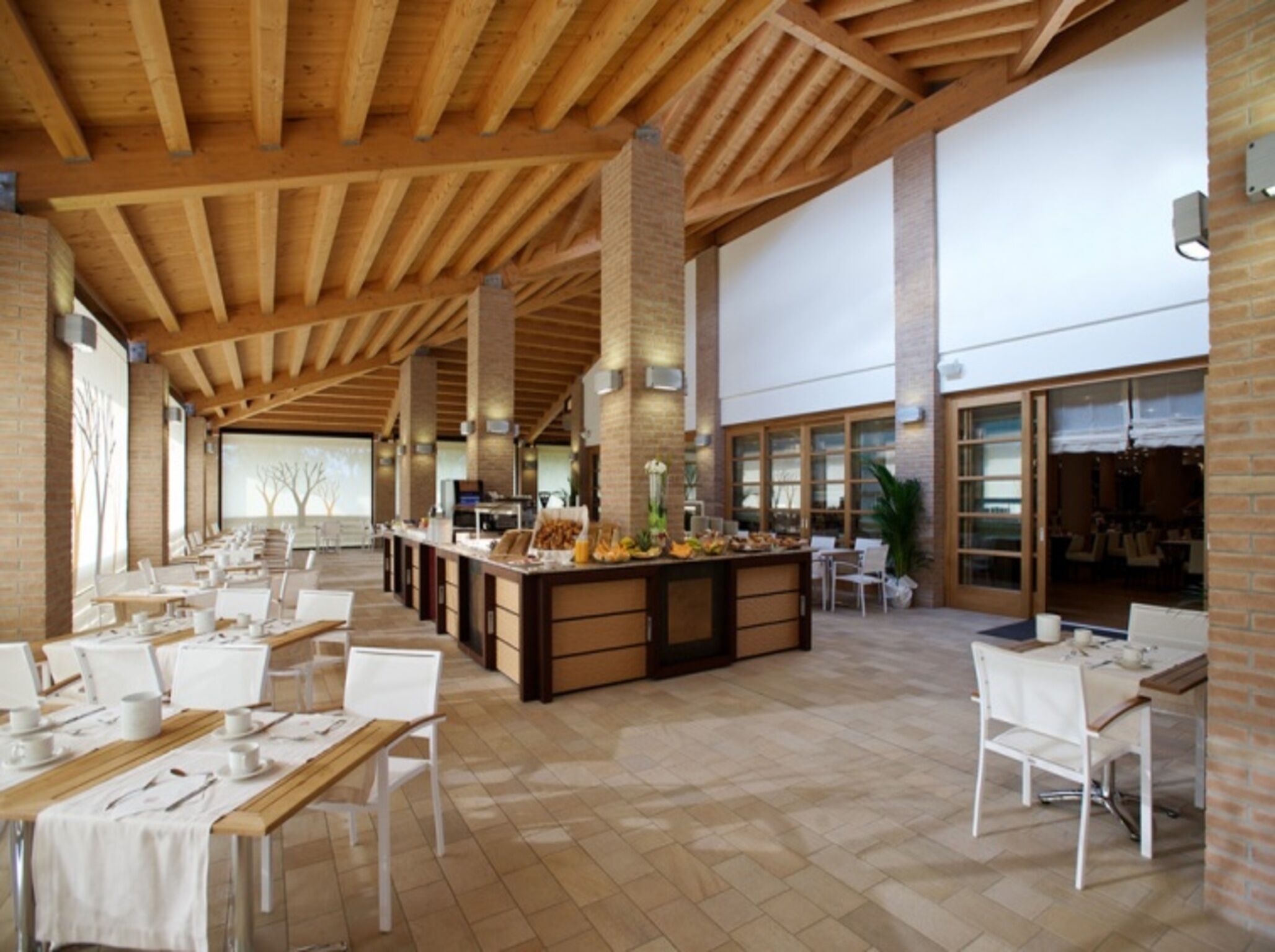 A spacious dining area with high ceilings and exposed wooden beams, featuring long tables set for dining and a central counter with food items.