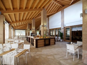 Un amplio comedor con techos altos y vigas de madera a la vista, con largas mesas dispuestas para comer y una barra central con alimentos.