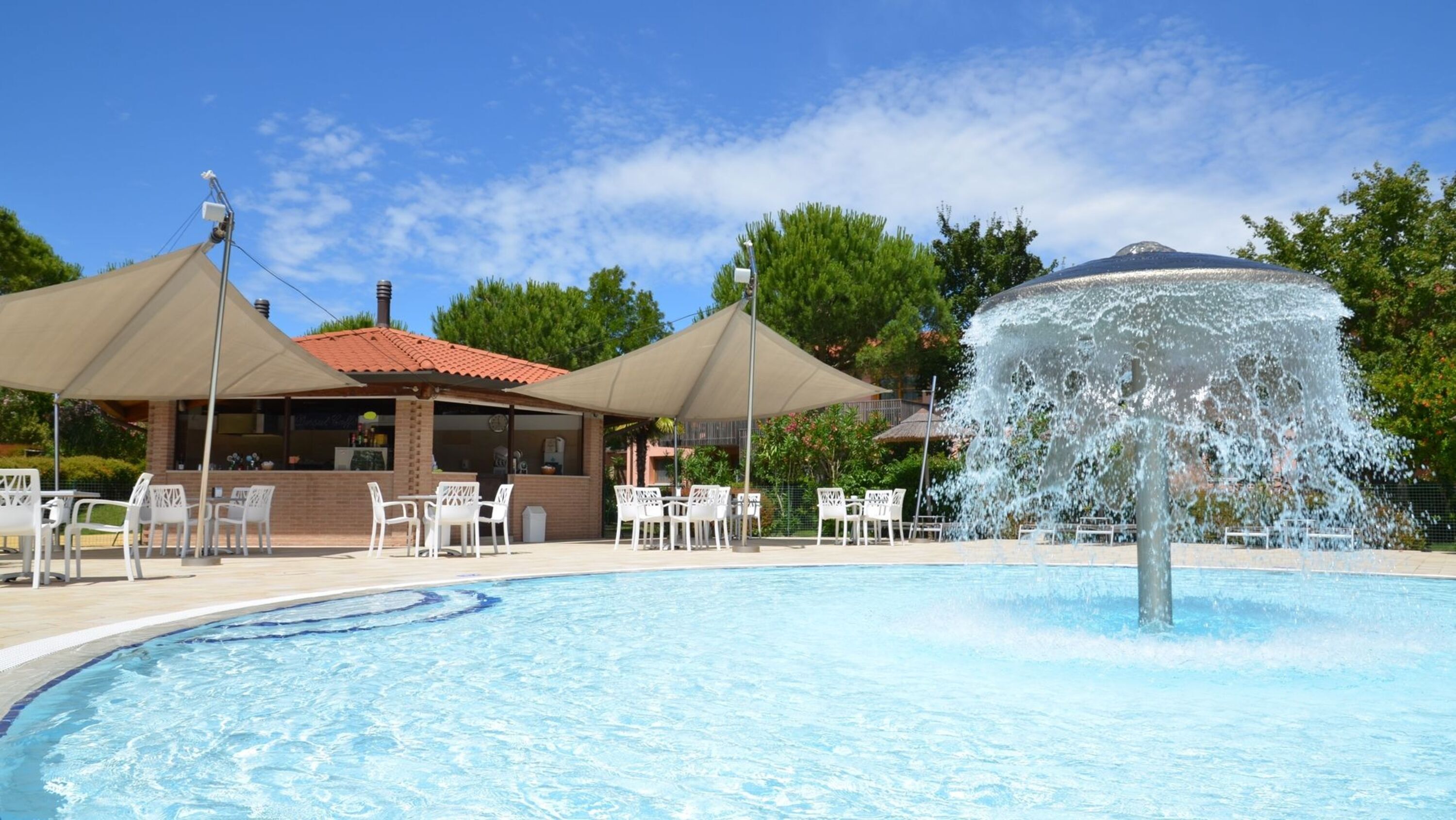 A swimming pool with a fountain, surrounded by white chairs and umbrellas, and a building with a red roof in the background.