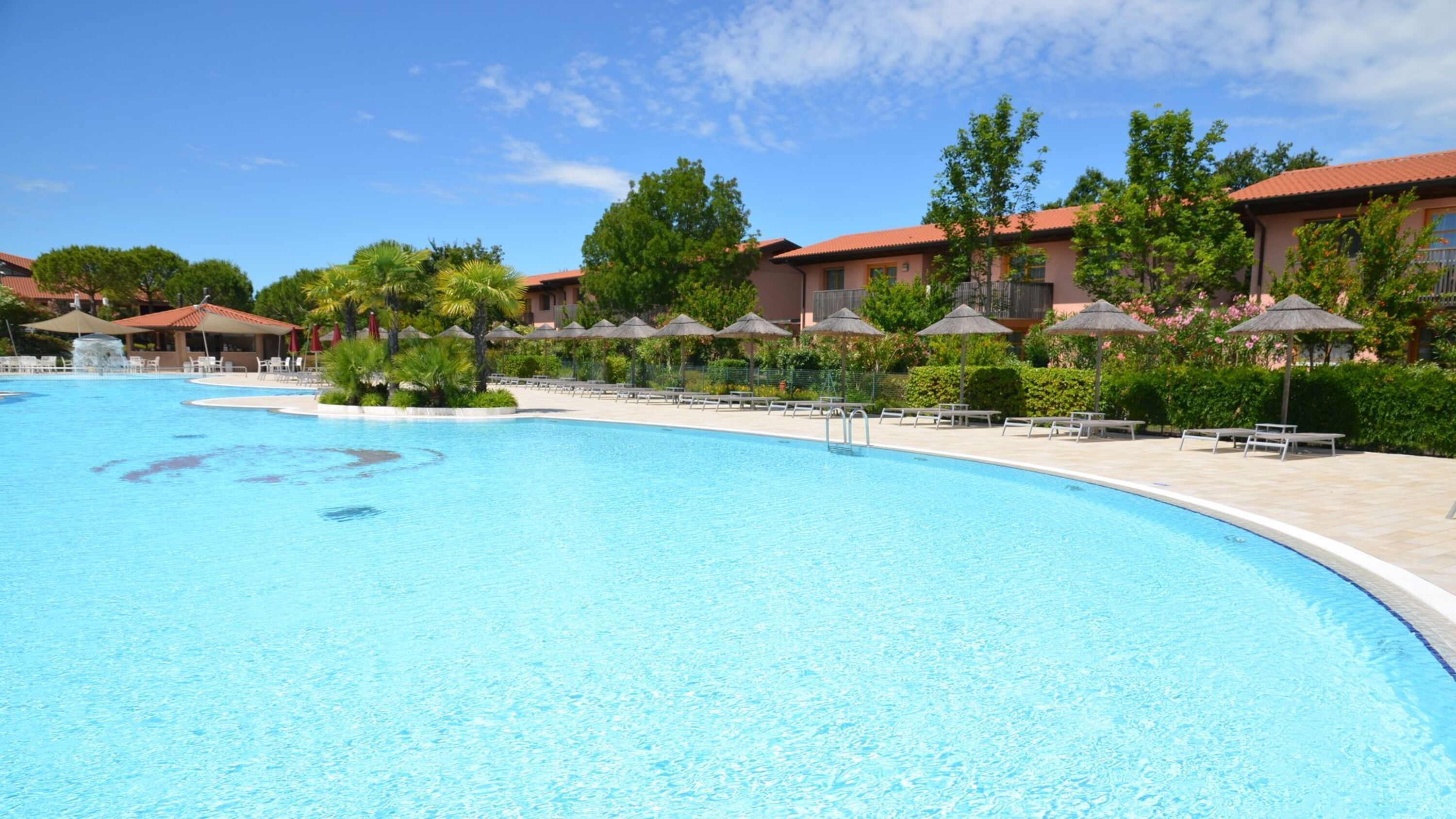 Une grande piscine, dotée de chaises longues et de parasols, est entourée de bâtiments aux toits rouges.
