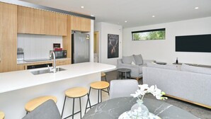 A modern kitchen with wooden cabinets, a white countertop, and a built-in microwave. There is a grey sofa and a dining table with stools in the living area. - Modern Comfort in 2-Bedroom Townhouse in Phillipstown (Christchurch)