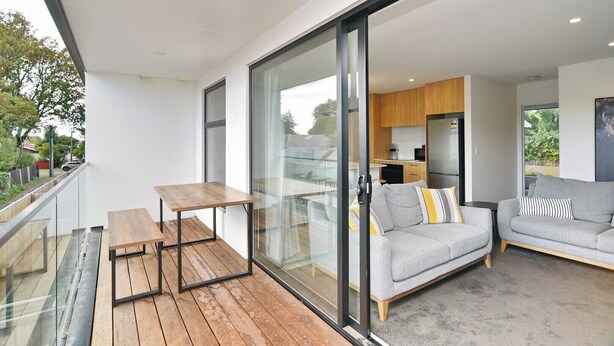 A modern balcony with a wooden table and a sofa, a glass door leading to a kitchen, and a view of the outdoors. - Modern Comfort in 2-Bedroom Townhouse in Phillipstown (Christchurch)