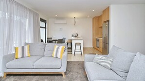 A modern living room with two grey sofas, a dining area with a small table and chairs, and a kitchen with a refrigerator and stove. - Modern Comfort in 2-Bedroom Townhouse in Phillipstown (Christchurch)