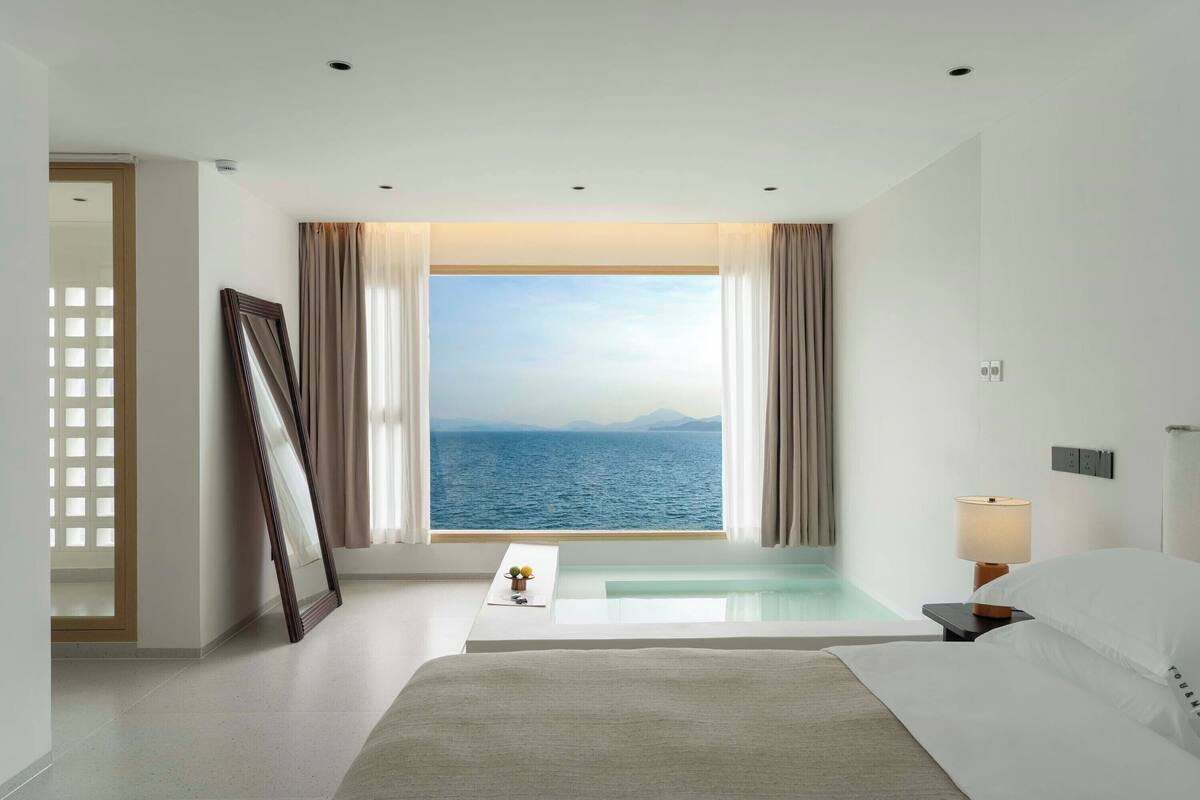 Una camera d'albergo con una grande finestra vista mare, un letto, un comodino con una lampada e un tavolino con un vaso di fiori.