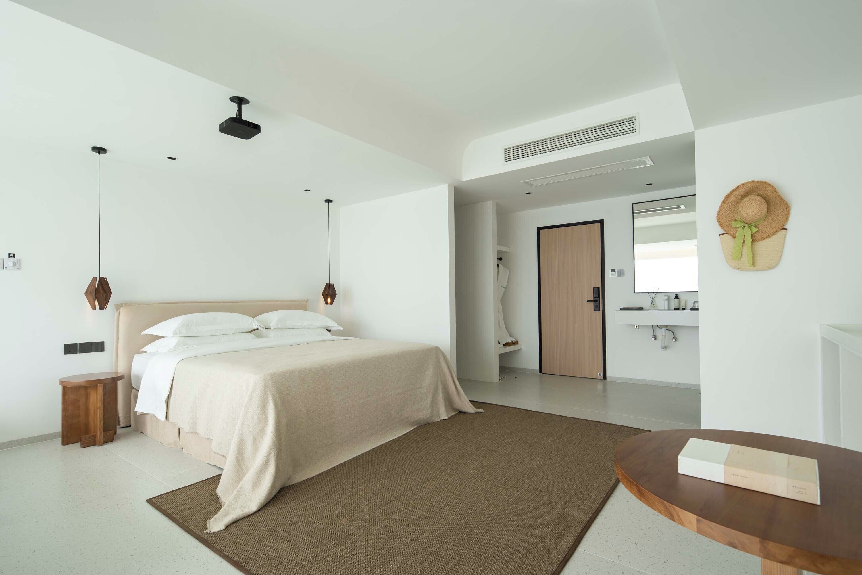 A modern bedroom with a large bed, bedside tables, a wooden nightstand, a round wooden table, and a wall-mounted hat.