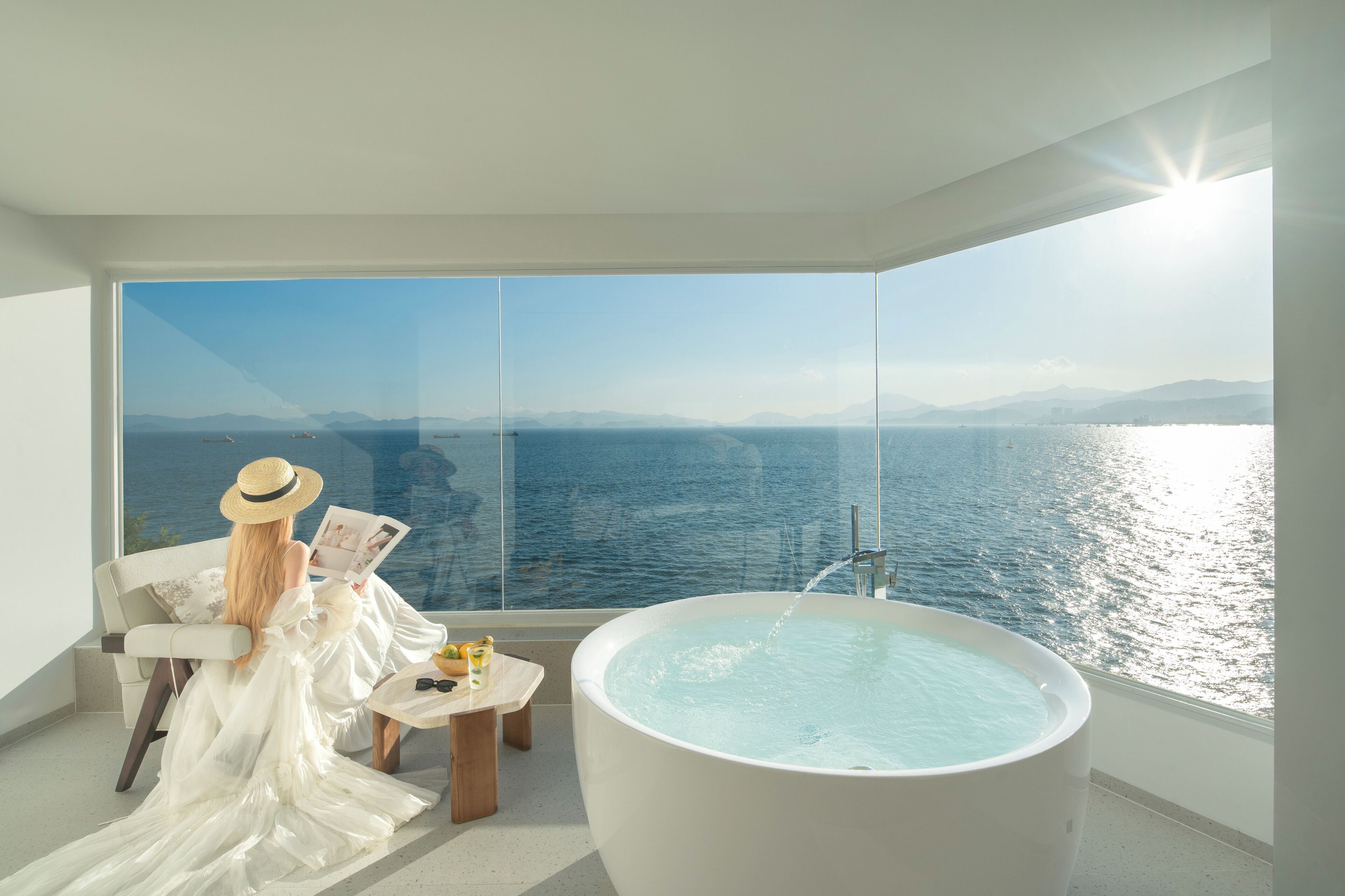 Une pièce avec une grande baignoire, une chaise et une table avec une boisson. Il y a une grande fenêtre qui offre une vue sur la mer et les montagnes.