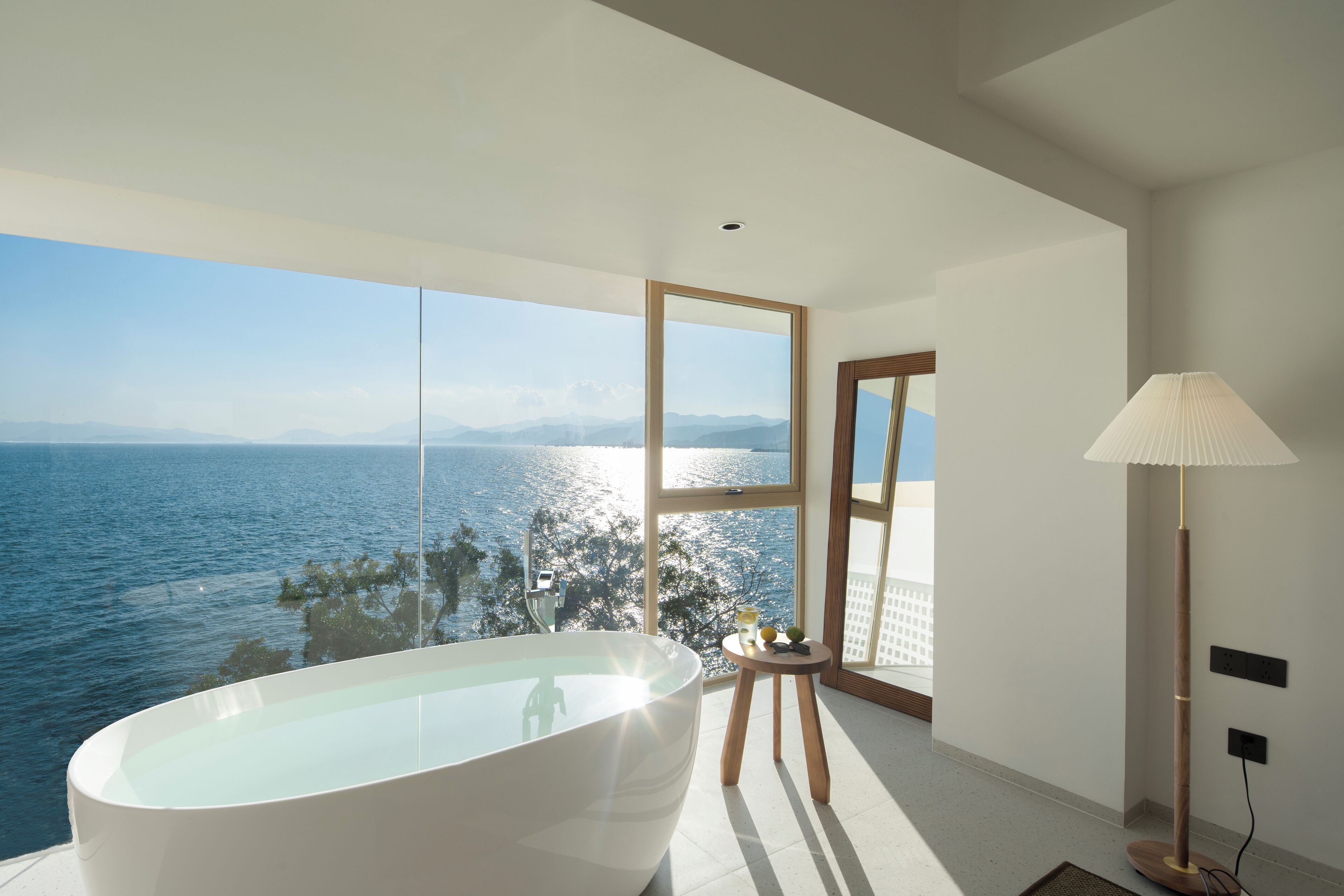 A modern bathroom with a freestanding bathtub, a large window offering a view of the sea, and a floor lamp.