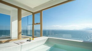 A modern bathroom with a large bathtub, a window with a view of the sea, and a small table with a book and glasses. - Shenzhen Secret Wild Reef Beautiful Stay (Shenzhen)