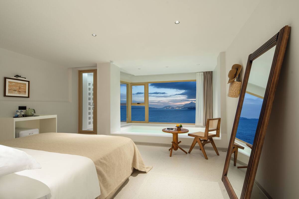 Una camera d'albergo con un letto grande, un tavolino rotondo con sedie, uno specchio ampio e vista mare da una grande finestra.