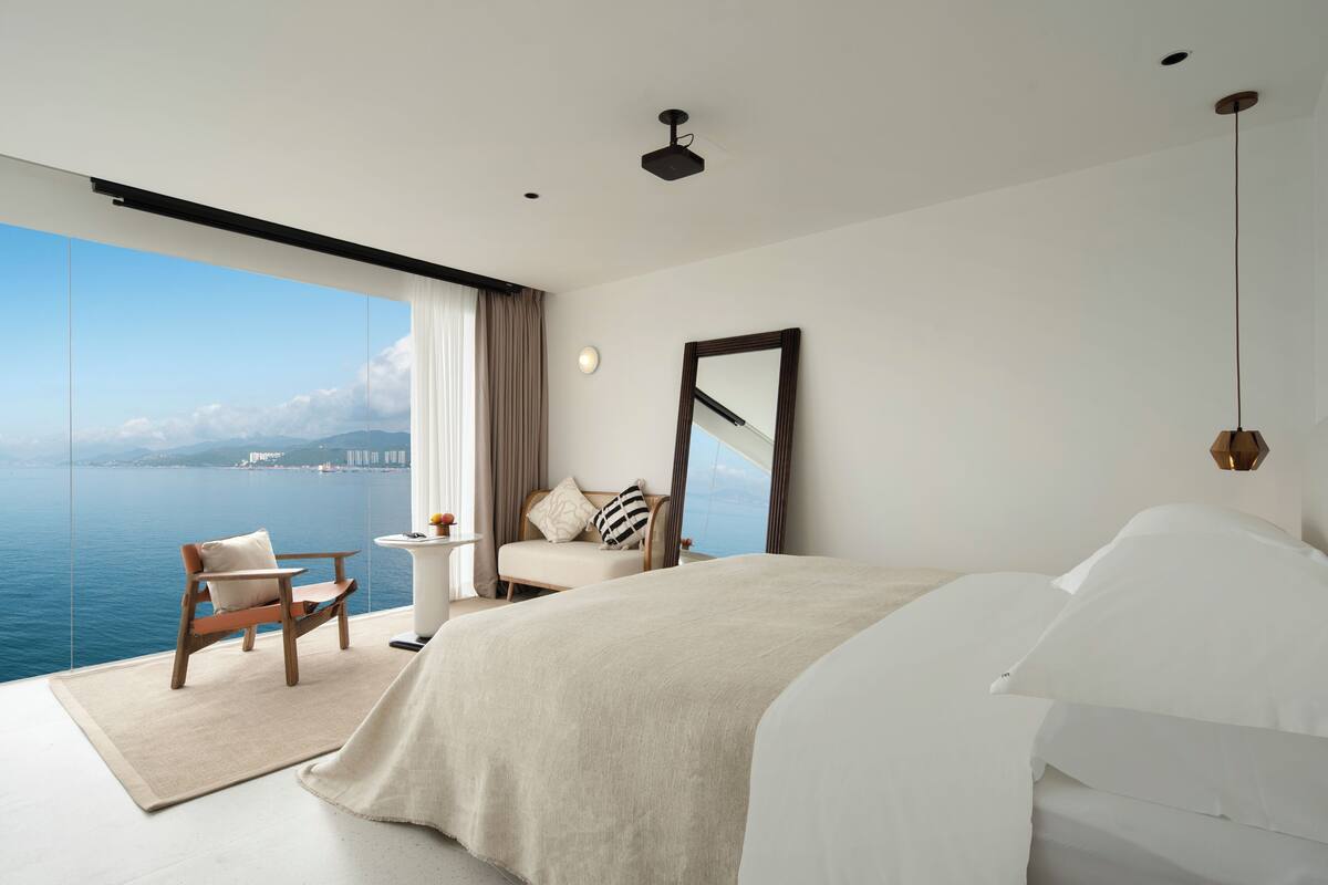Una camera da letto moderna con un grande letto, vista mare, una sedia e un tavolino.