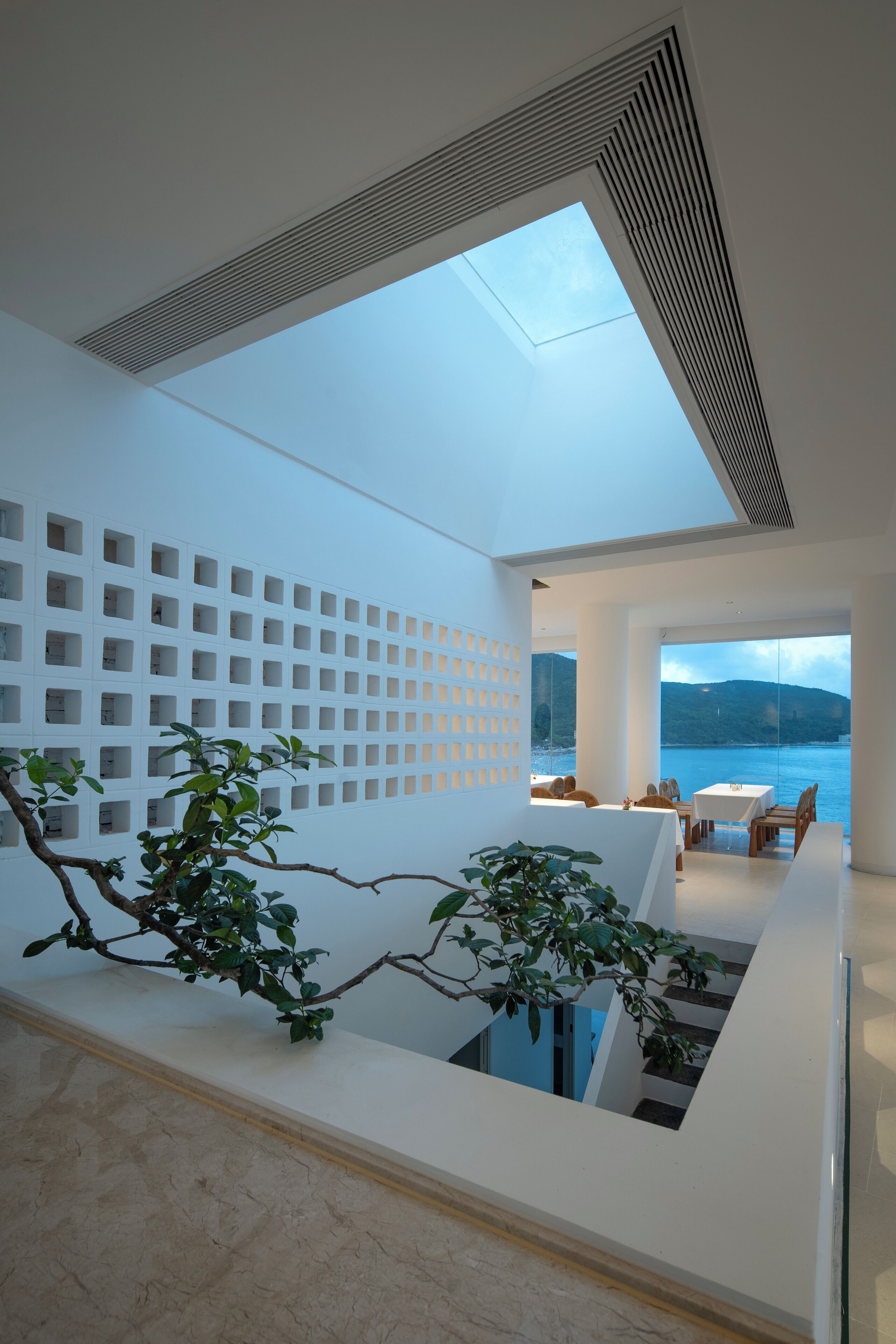 A modern interior with a skylight, a white wall with square cutouts, a dining area with tables and chairs, and a view of the sea.