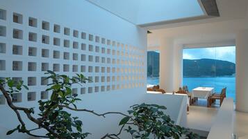 A modern interior with a skylight, a white wall with square cutouts, a dining area with tables and chairs, and a view of the sea.