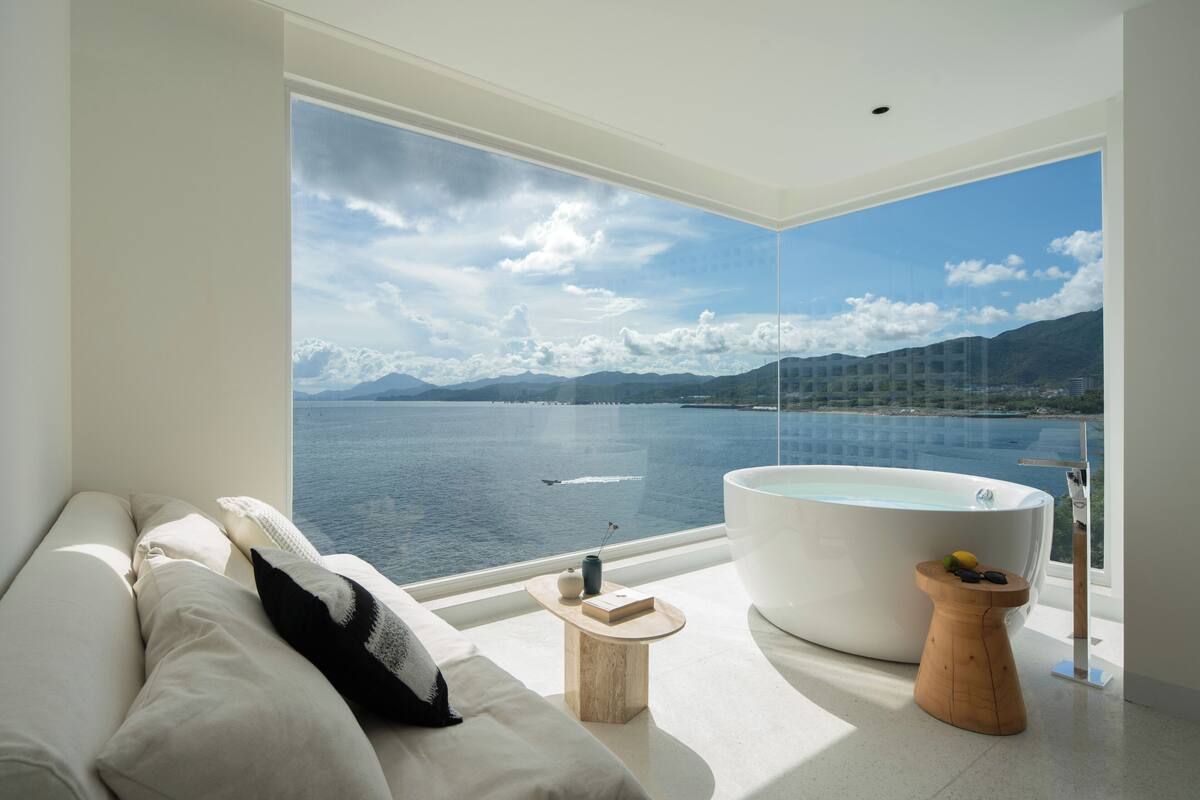 Una camera moderna con una grande finestra che si affaccia su un lago e montagne. C'è un divano, un tavolino rotondo in legno e una vasca da bagno ampia.