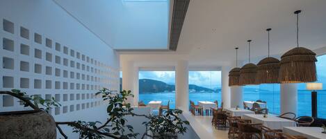 A modern dining area with a view of the sea, white walls, and natural light from a skylight.