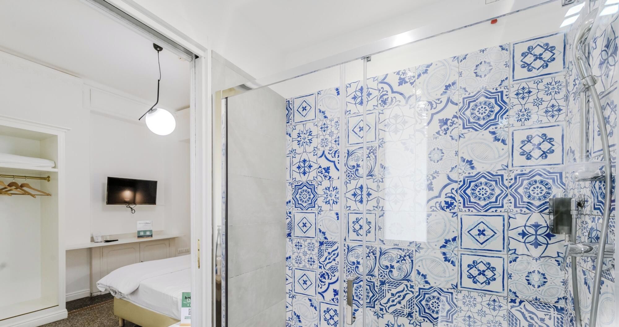 A hotel room with a bed, a TV, a shower with blue patterned tiles, and a hanging light fixture.