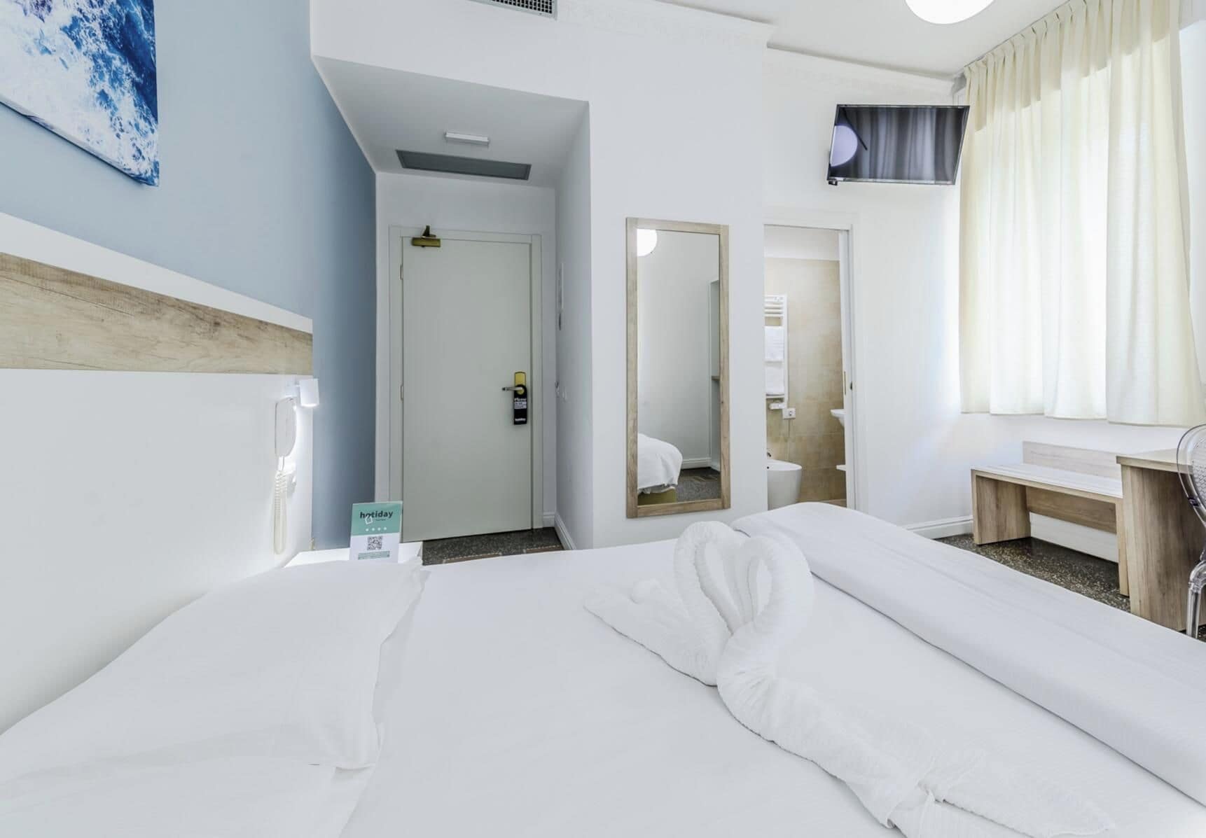 A hotel room with a bed, a desk, a mirror, and a bathroom visible through an open door.